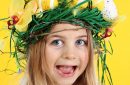 Young happy girl wearing straw headdress made of spring flowers, Easter eggs and feathers on yellow background.