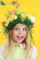 Young happy girl wearing straw headdress made of spring flowers, Easter eggs and feathers on yellow background.