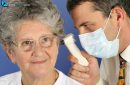 A doctor with a mask looks into the patient's ear with an otoscope. The patient is a woman over 65